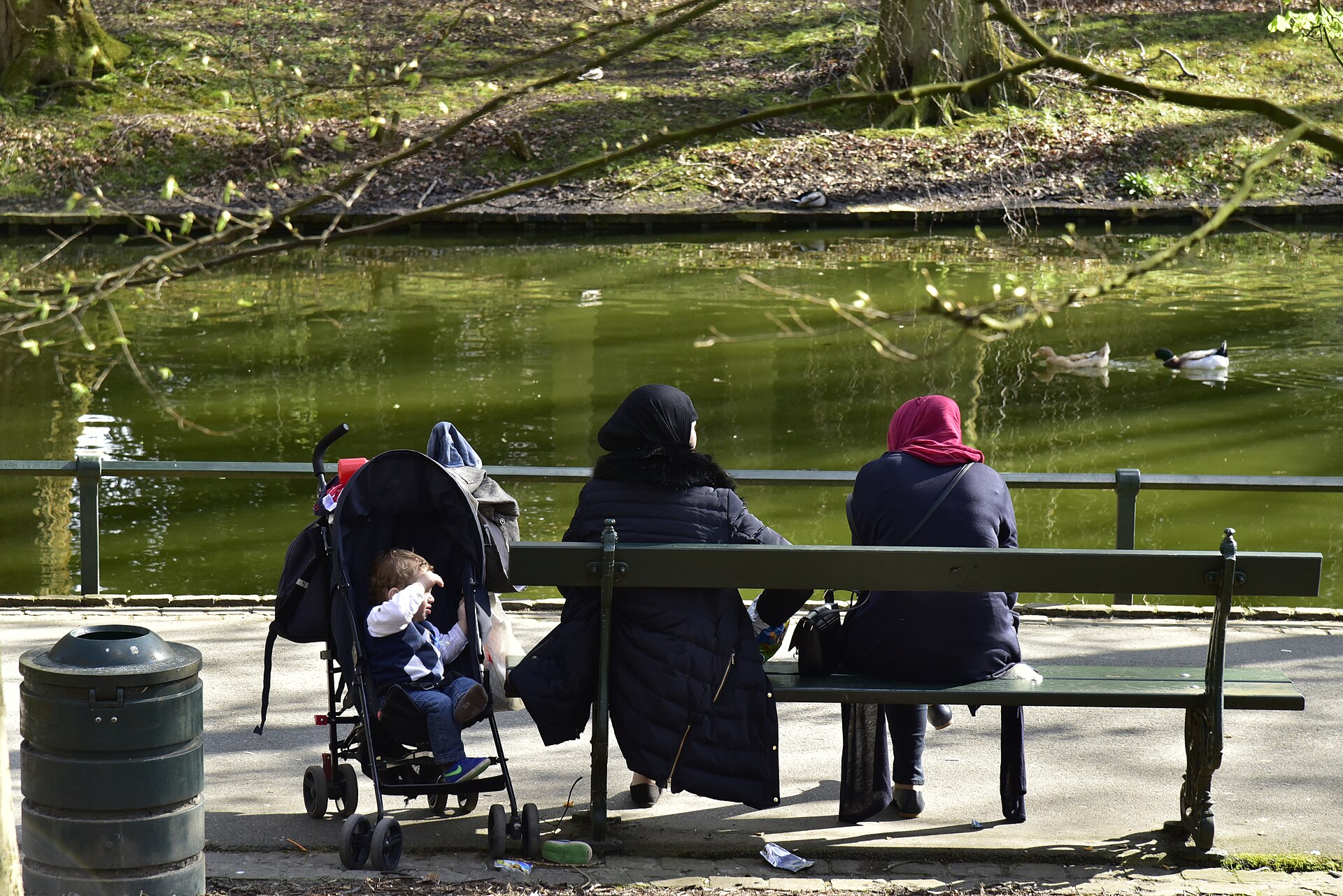 Two adults sit beside a stroller on a pondside bench in Brussels’ Josaphat Park, watching ducks on the water.