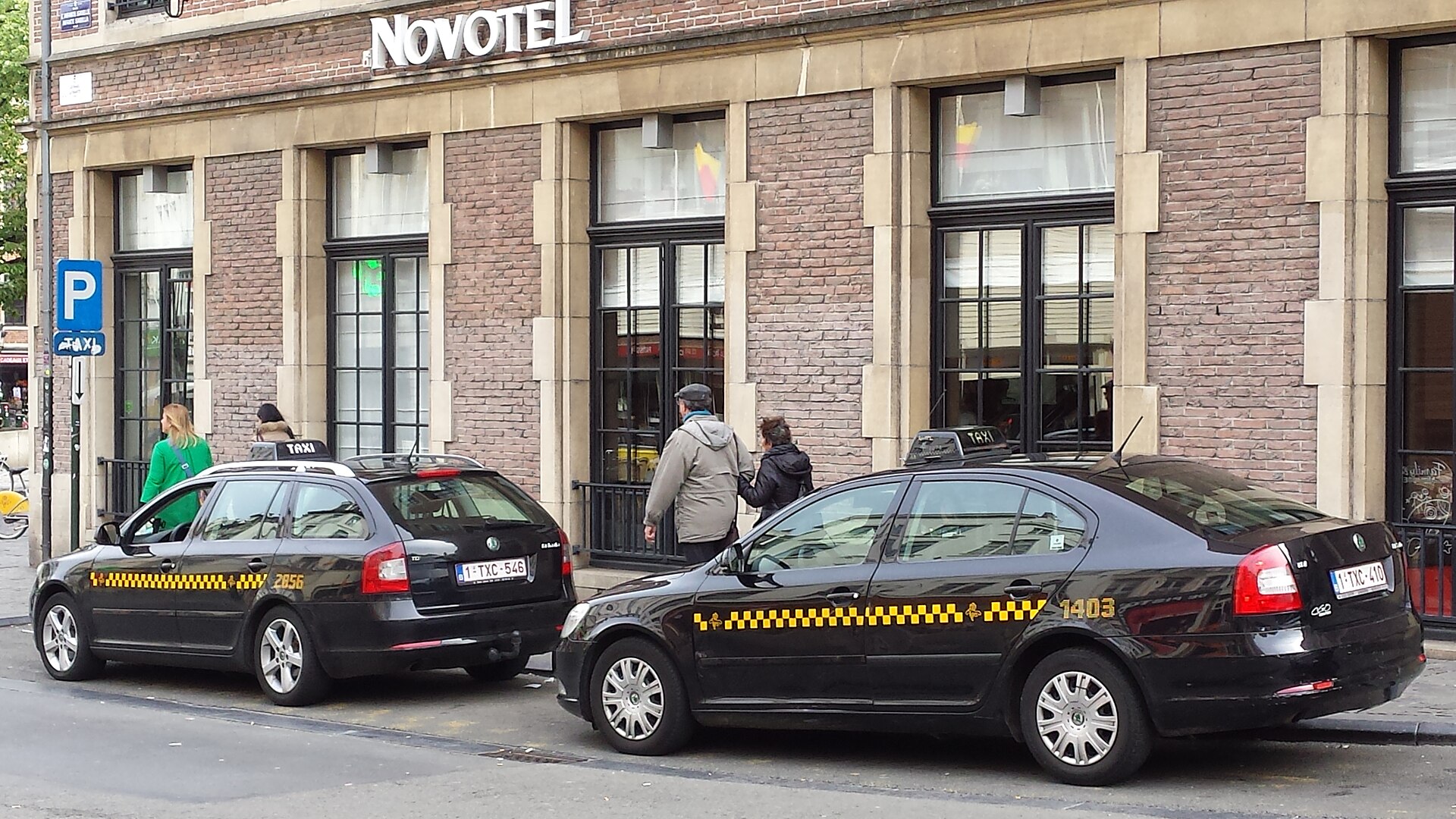 Two black Brussels taxis with yellow checker stripes and roof signs wait beside a marked taxi stand outside a Novotel.