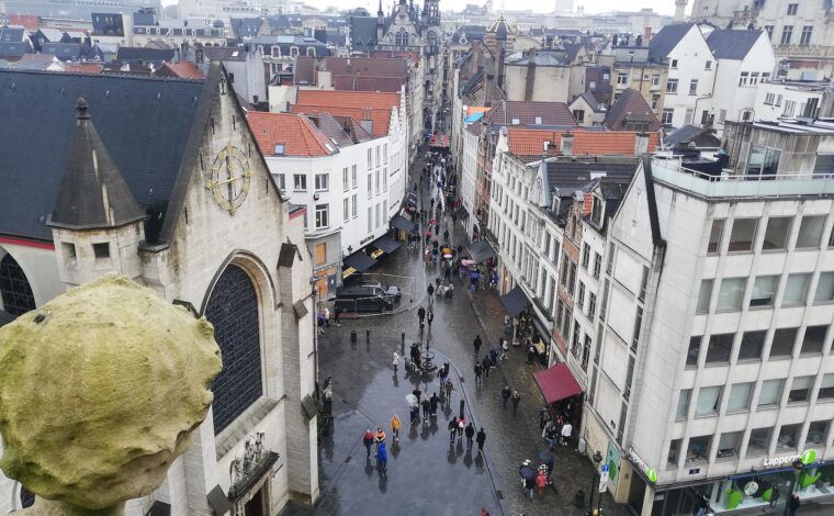 Umbrellas dot wet Rue au Beurre in central Brussels, with Saint Nicholas Church beside the street and historic rooftops beyond.