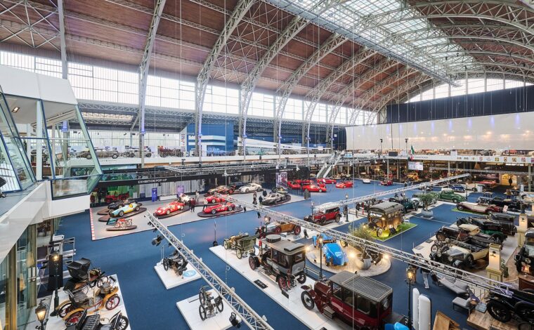 Vintage cars and sports cars fill Autoworld in Brussels beneath a soaring iron roof, viewed from the upper gallery.