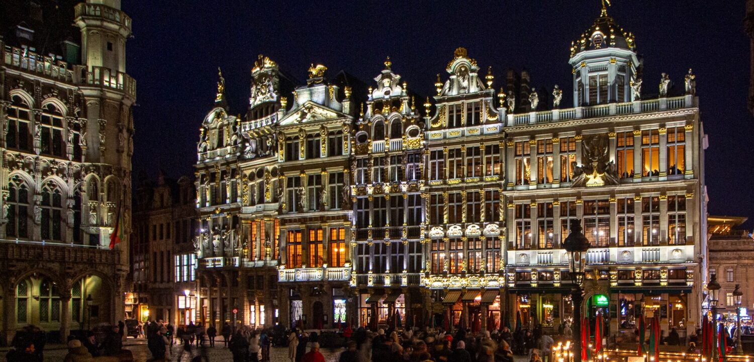 Visitors cross Grand-Place in Brussels at night beneath illuminated guildhouse facades, with part of the Gothic Breadhouse visible at left.