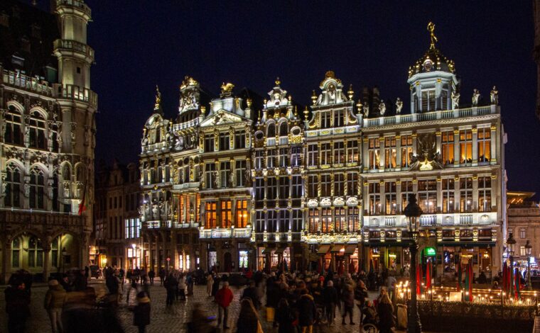 Visitors cross Grand-Place in Brussels at night beneath illuminated guildhouse facades, with part of the Gothic Breadhouse visible at left.