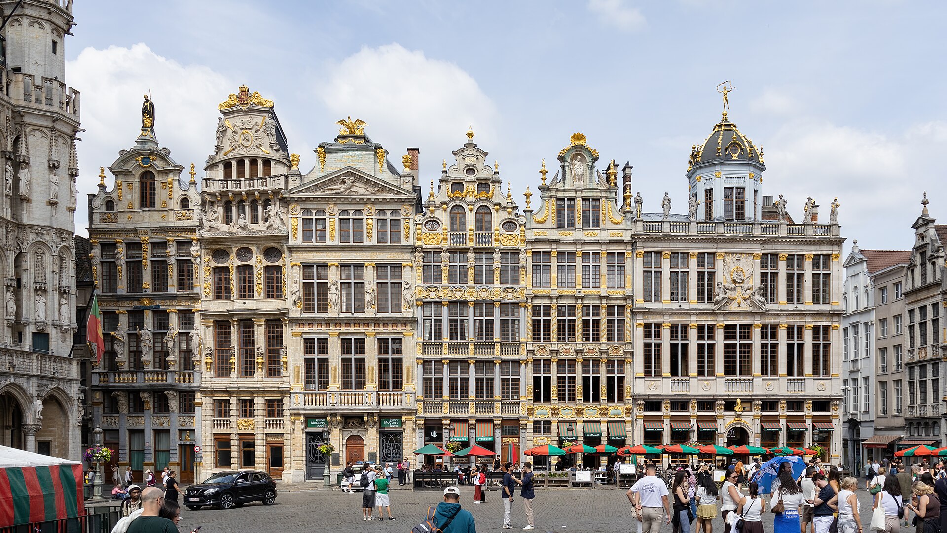 Visitors gather below ornate guildhouses on the Grand-Place in Brussels, with gilded statues, cafe umbrellas and cobblestones visible.