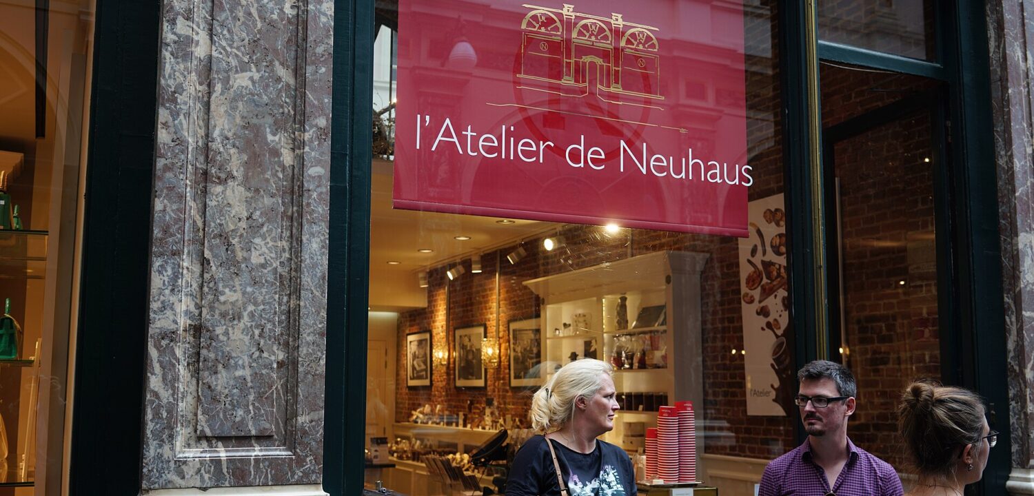 Visitors stand outside Atelier de Neuhaus in Brussels, beneath a red sign beside the illuminated chocolate shop window.