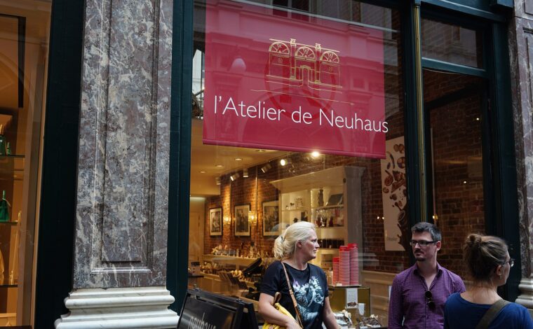 Visitors stand outside Atelier de Neuhaus in Brussels, beneath a red sign beside the illuminated chocolate shop window.