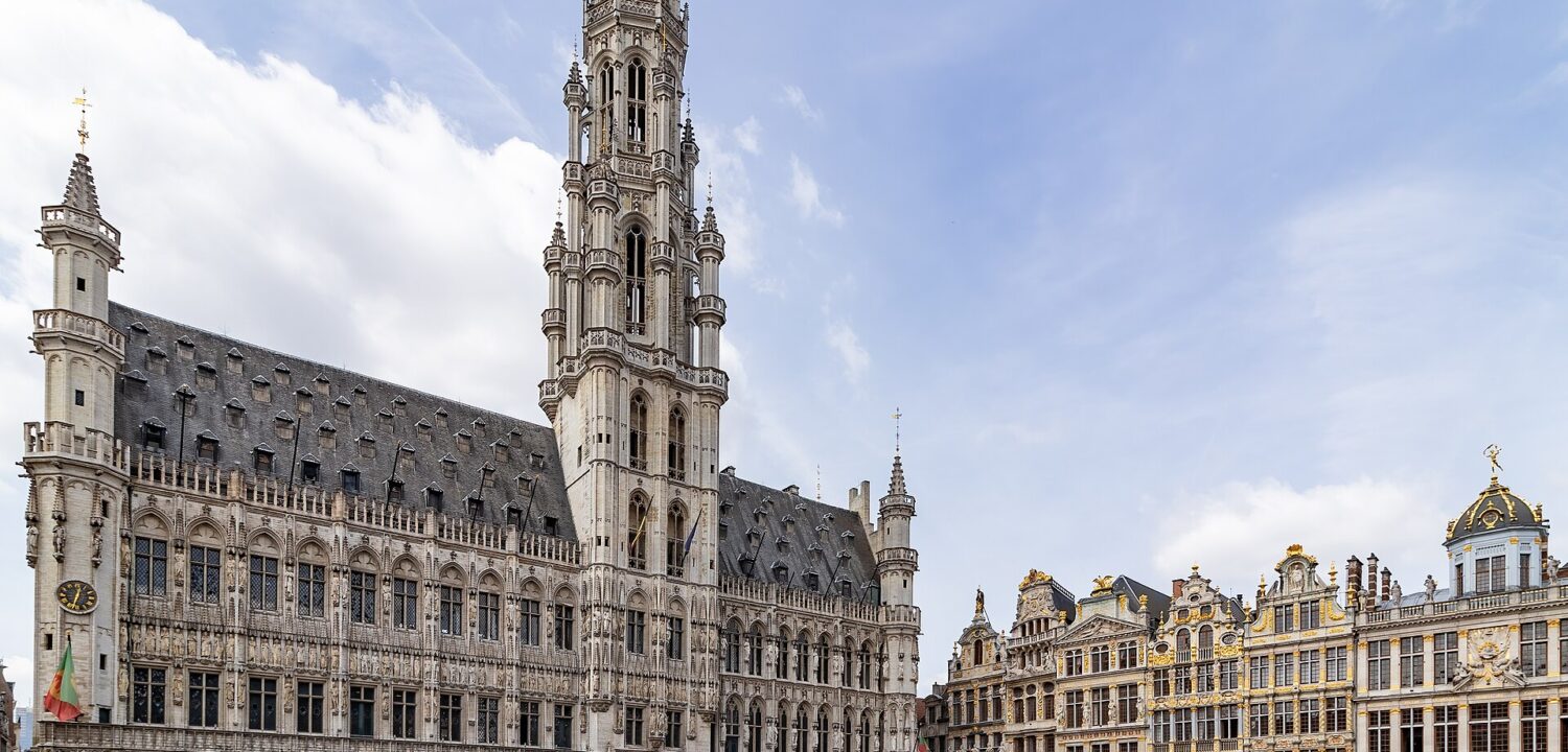 Visitors stroll across Brussels’ Grand-Place beneath the Town Hall’s Gothic tower and ornate gilded guildhouses on a summer afternoon.