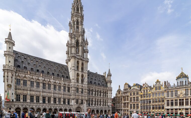 Visitors stroll across Brussels’ Grand-Place beneath the Town Hall’s Gothic tower and ornate gilded guildhouses on a summer afternoon.