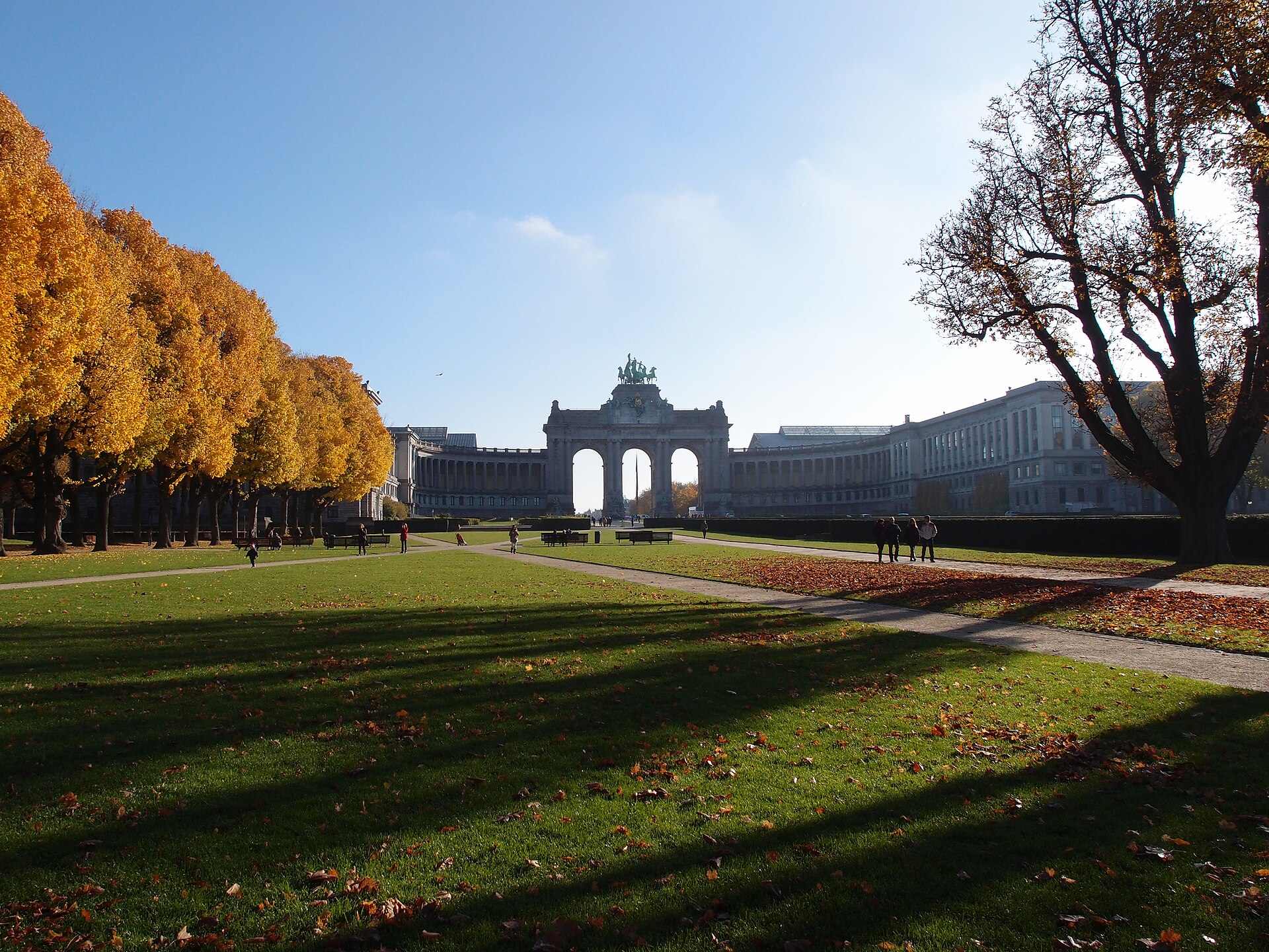 Visitors stroll along paths through autumn lawns in Brussels' Cinquantenaire Park, with benches, golden trees and the triumphal arcade beyond.