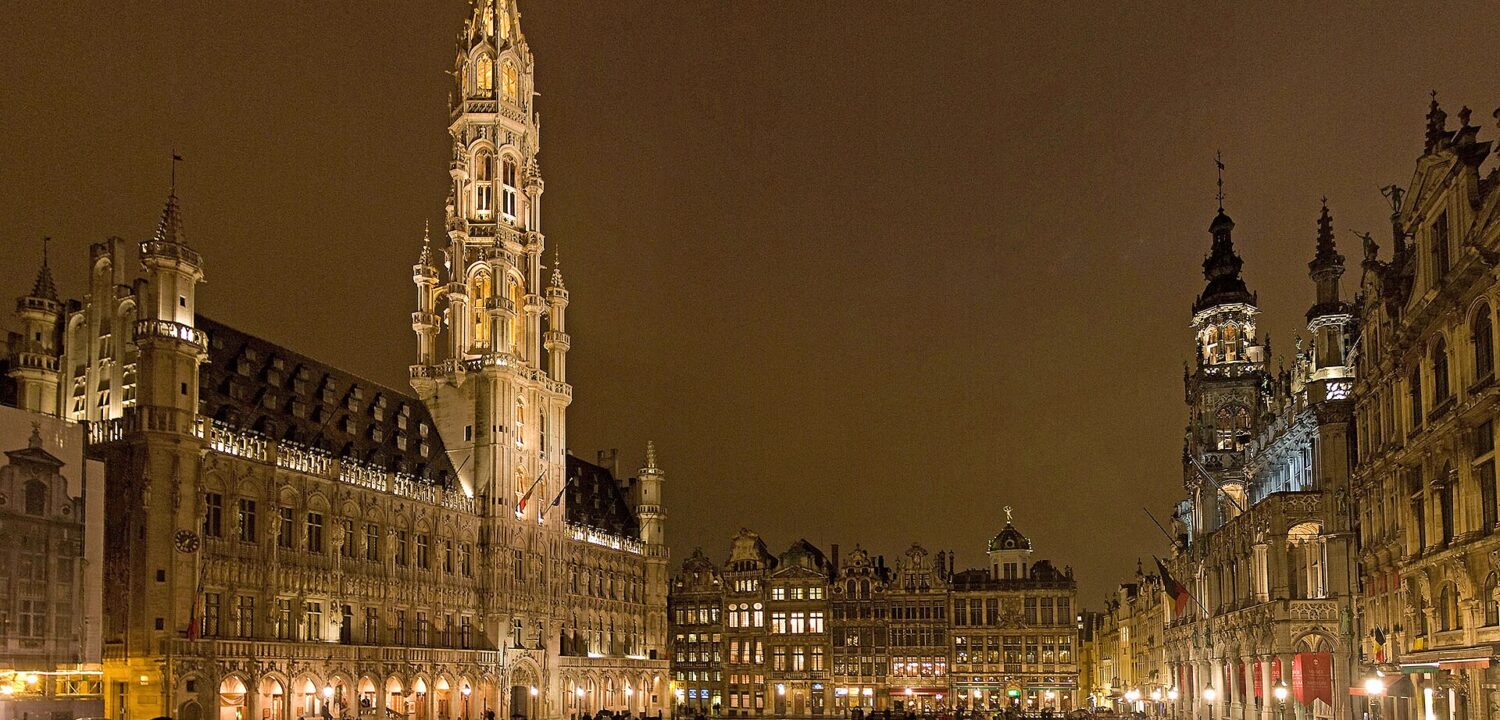 Visitors with umbrellas cross rain-soaked Grand-Place in Brussels beneath the illuminated town hall and surrounding guildhouses at night.