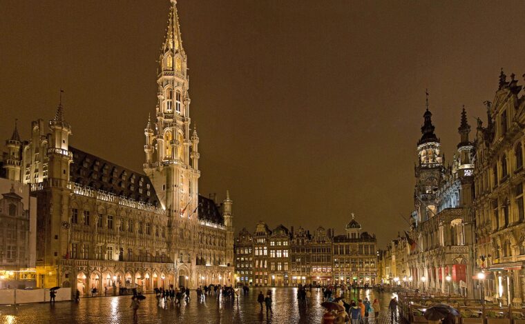 Visitors with umbrellas cross rain-soaked Grand-Place in Brussels beneath the illuminated town hall and surrounding guildhouses at night.