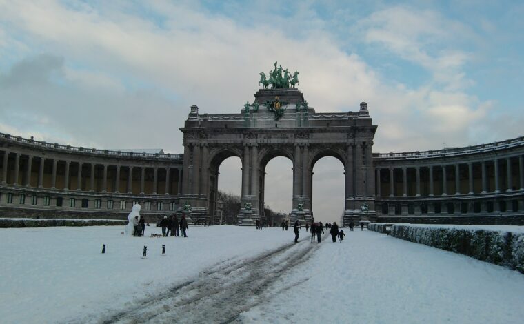 Visitors walk through snow toward the triple arch in Brussels' Cinquantenaire Park, with a snowman standing beside the path.