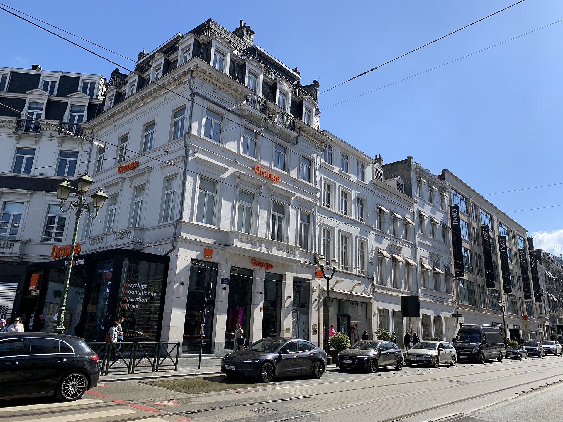 White facades and INNO storefronts line the entrance to Avenue Louise in Brussels, beside tram tracks, cars and pedestrians in daylight.