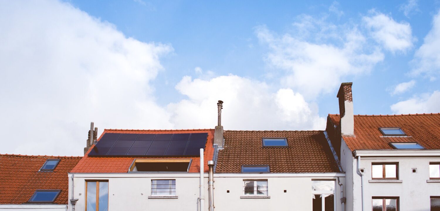 White residential facades in Brussels have tall windows, red tiled roofs, skylights and solar panels beneath a blue, clouded sky.