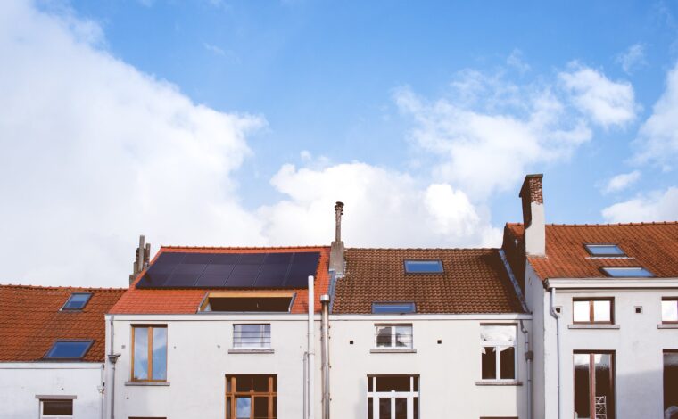 White residential facades in Brussels have tall windows, red tiled roofs, skylights and solar panels beneath a blue, clouded sky.
