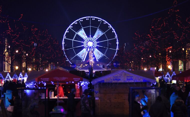 Wooden chalets and visitors fill Brussels Winter Wonders beneath red-lit trees, with the illuminated Ferris wheel rising behind the market.