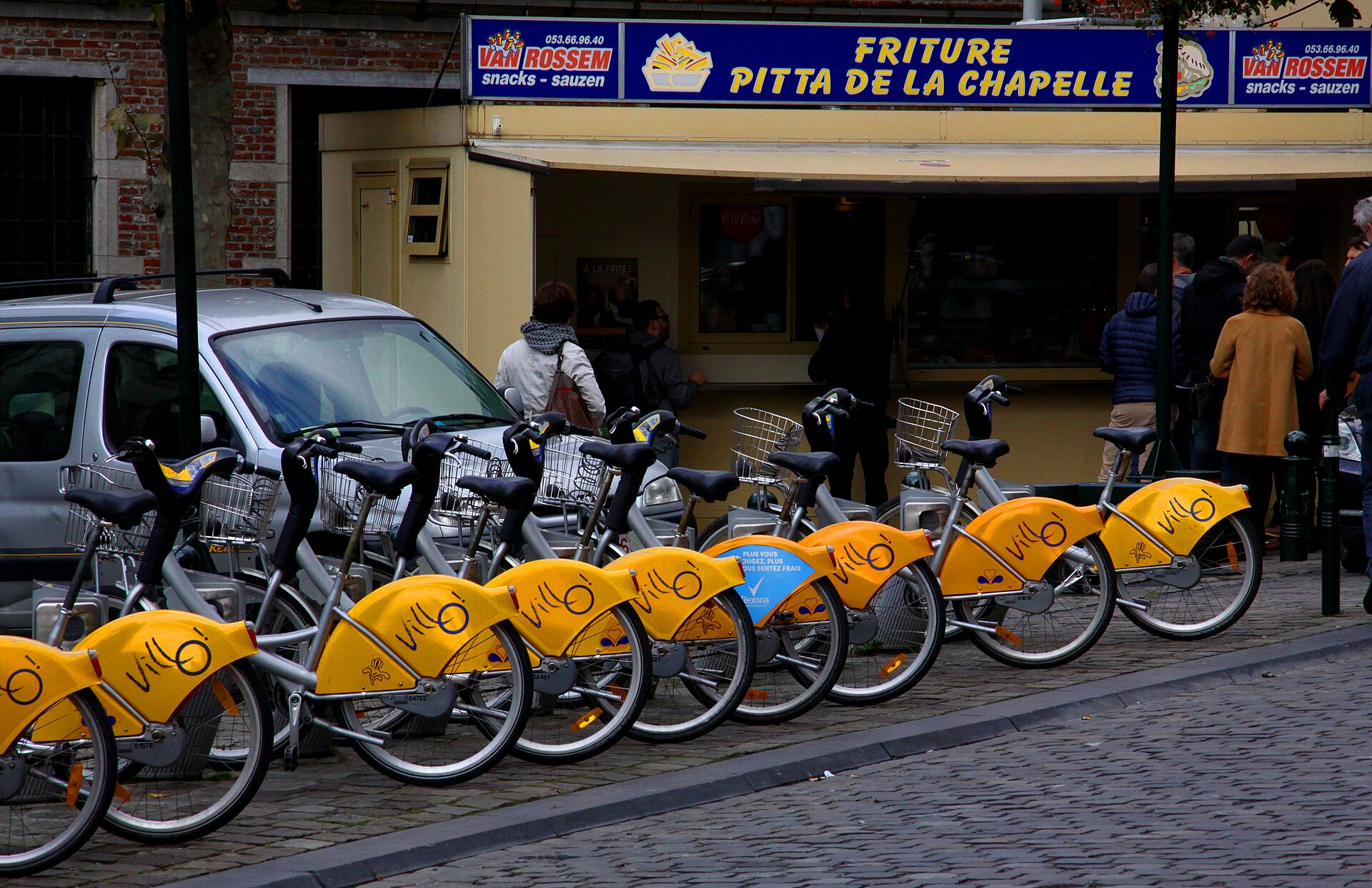 Yellow Villo! shared bicycles stand docked outside a snack shop in the Marolles neighbourhood of Brussels, with customers behind.