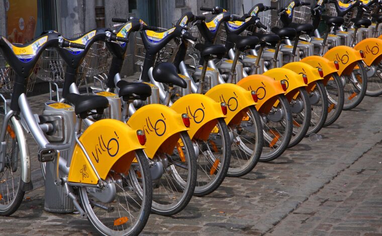 Yellow Villo! wheel guards line a row of silver shared bicycles docked on a cobbled street in central Brussels.