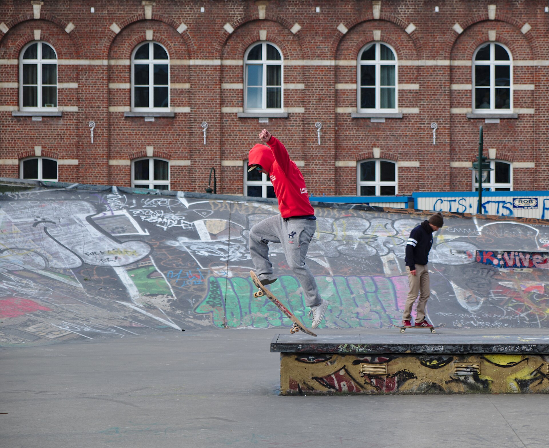 Young skateboarders practise tricks beside a graffiti-covered bowl at Skatepark des Ursulines in Brussels, with a brick building behind them.